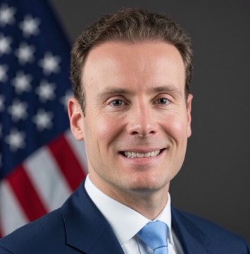 A professional headshot of a smiling man in a dark suit and light blue tie, posed in front of an American flag—identified by the accompanying post as CFTC Chair Mike Selig, who is announcing a shift toward crypto-friendly regulation.
