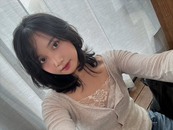 A selfie of a young woman with short dark hair, wearing a beige cardigan over a lace top, smiling softly near a window with sheer curtains.