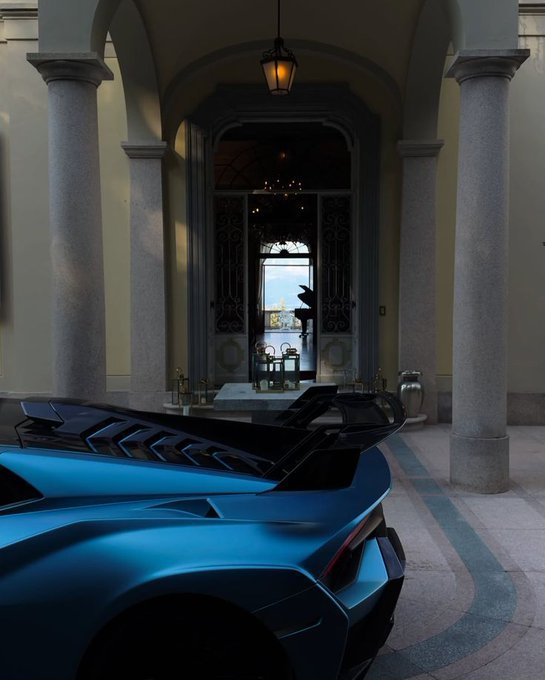 The rear of a blue sports car with a prominent spoiler is parked in a grand, columned entryway, looking through to a courtyard beyond.