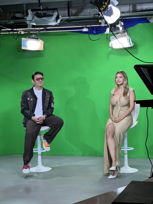 Two people, a man in a dark jacket and sunglasses and a woman in a sparkly gold dress, sit on stools in a studio in front of a green screen with professional lighting equipment visible above.