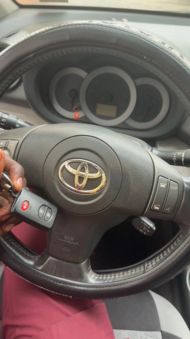 A person holds a new Toyota key fob with a red panic button in front of the steering wheel of a Toyota vehicle, featuring the Toyota logo. The image illustrates the post about getting a new replacement key for the car.