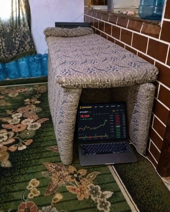 A laptop displaying the Binance BTC/USDT chart with Bitcoin at 78,000 sits on a patterned floral rug beneath a cushioned stool, perfectly tucked away as a quiet spot to monitor the crypto market.