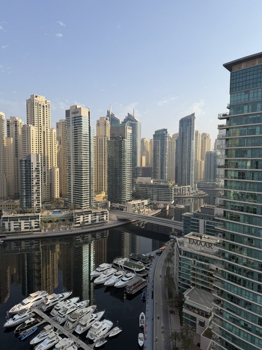 An aerial view of Dubai Marina showing numerous yachts docked in the harbor, surrounded by tall skyscrapers including one labeled 'EMAAR'.