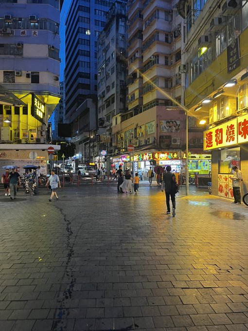 An evening street scene in Hong Kong with illuminated yellow shop signs, pedestrians walking on a wet tiled street, and tall residential buildings.