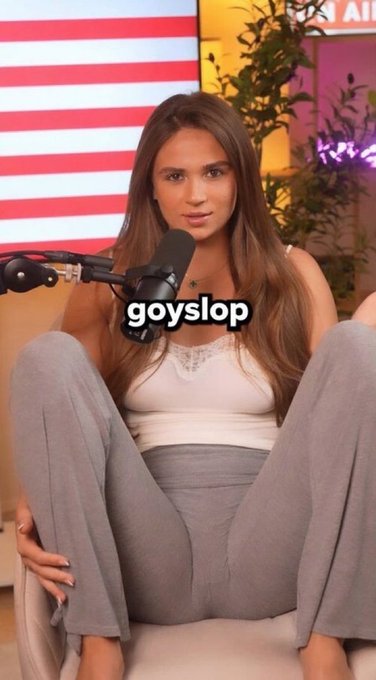 A young woman with long brown hair sits cross-legged in front of a microphone, with the text "goyslop" overlaid across her chest and an American flag visible in the background. The image relates to the post suggesting she would have strong engagement promoting memecoins, with the "goyslop" text adding a meme-style overlay.