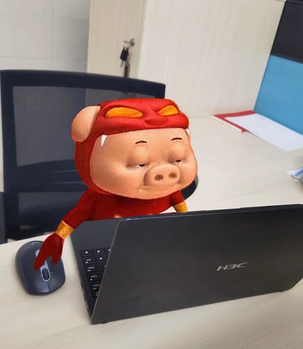 A plush toy of Zhu Bajie (the pig character from Journey to the West) in a red superhero costume sitting on an office desk in front of an open laptop, appearing to work or browse.