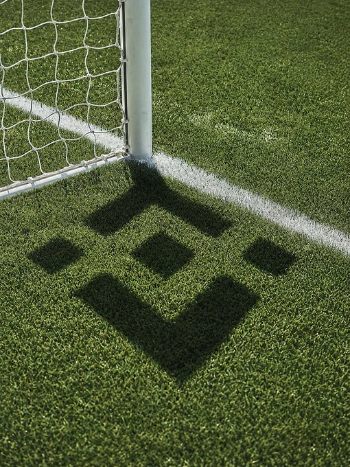 A close-up of a soccer field goalpost casting a distinctive shadow on the artificial turf near the white sideline.