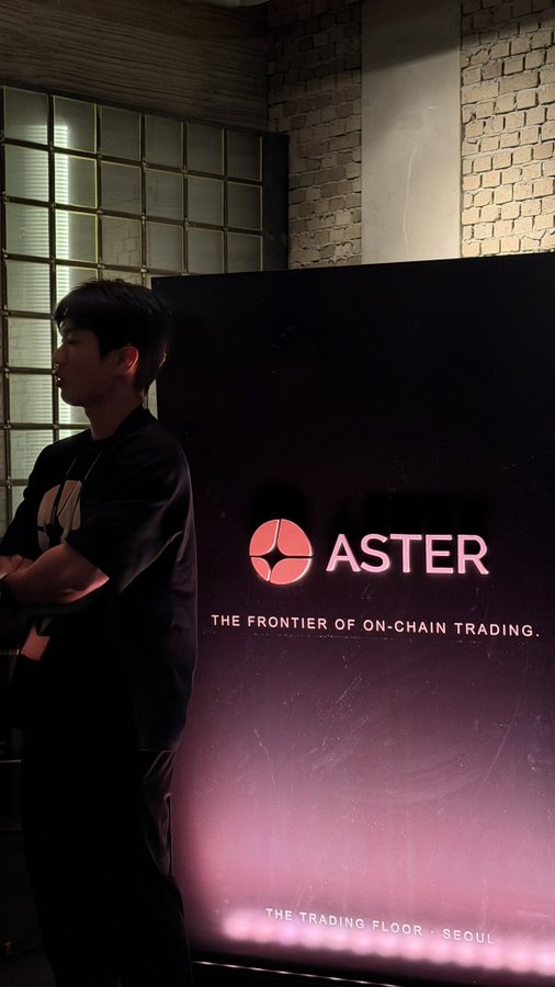 A young man stands with his arms crossed next to a glowing sign that reads 'ASTER - THE FRONTIER OF ON-CHAIN TRADING - THE TRADING FLOOR, SEOUL'.