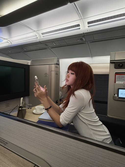 A woman with long auburn hair sits in an airplane's premium suite, taking a selfie on her phone with a coffee cup on the tray table, while a label reading "A350-900H" is visible on the seat console—aligning with the post's mention of attending Token2049 last minute to film content with brand partnerships.