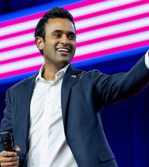 A man in a dark suit and white shirt smiles broadly while pointing, standing in front of an American flag backdrop.