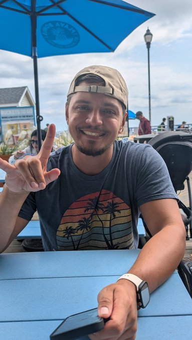 A smiling young man wearing a backwards tan baseball cap and a t-shirt with a sunset and palm tree graphic, sitting at a blue outdoor table under a blue umbrella while making a hand gesture.