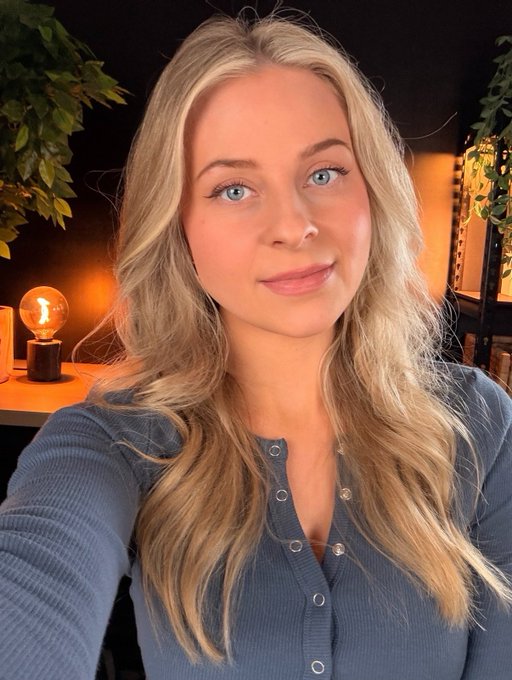 A selfie of a blonde woman in a blue button-up top, posing in front of warm ambient lighting and greenery. The image complements the post's message about steadfast loyalty to Cardano ($ADA), as her calm, confident gaze visually embodies the "not leaving" sentiment.