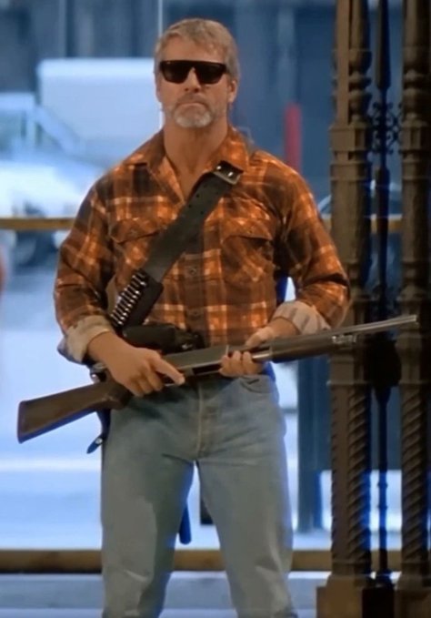 A man with a gray goatee wearing sunglasses, an orange plaid shirt, and jeans stands holding a pump-action shotgun with a bandolier across his chest in an urban setting.