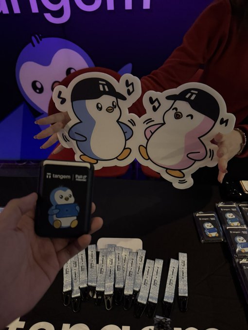 A vendor's hands hold up cute penguin character standees for 'tangem NF', with merchandise and a wallet displayed on the table.