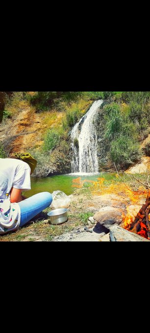 A person in a hat sits by a green pool at the base of a small waterfall, preparing food with a pot and kettle while a campfire burns nearby.