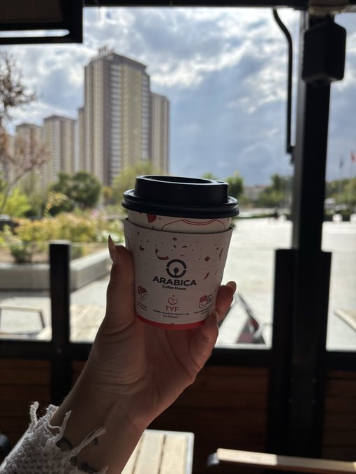 A hand holding a white paper coffee cup labeled 'ARABICA Coffee House' and 'TVP', with a city skyline and cloudy sky visible through a window in the background.