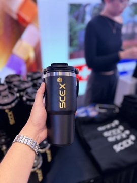 A hand holds a black tumbler branded with the "SCEX" logo at the RWA Hanoi Conference, with event materials and a woman in the background, visually tying into the post's news about @scexofficial's app transition, referral system, and multi-level commission structure updates.