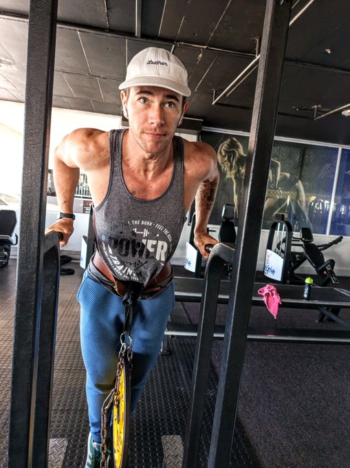 A woman in a gym wearing a white cap, grey 'POWER' tank top, and blue leggings performing a tricep dip on parallel bars using a weight belt and yellow weight plates.