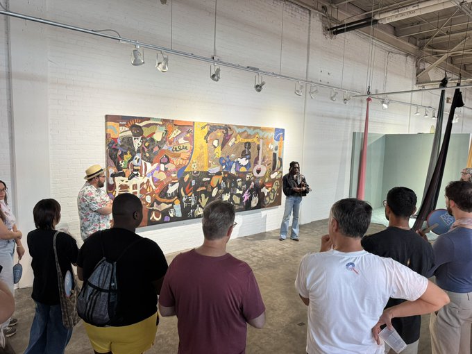 A group of people gathered in a white-walled art gallery listening to someone presenting in front of a large, colorful abstract mural painting.