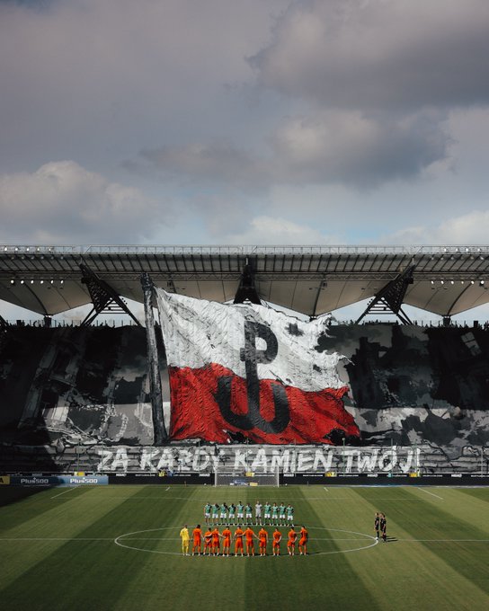 Two soccer teams line up on the field in a stadium where fans display a large tifo featuring the Polish flag colors and 'PU' logo, with a banner reading 'ZA KARDY KAMIEN TWOJ'.