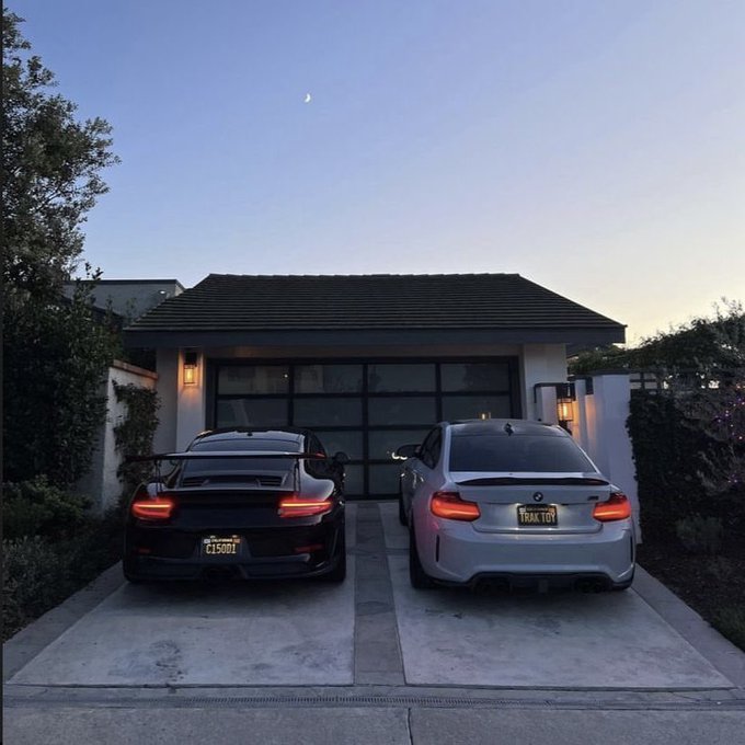 Two sports cars parked in a driveway at dusk: a black Porsche (license plate C15001) and a silver BMW M3 (license plate TRAX TOY) in front of a garage.