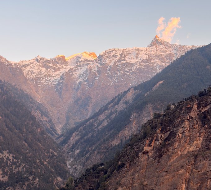 A scenic mountain landscape showing snow-capped peaks illuminated by golden sunlight, with a deep forested valley below and rocky cliffs on the right.
