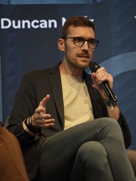 Duncan Moir of 21Shares speaks into a microphone while seated onstage, with partial backdrop text reading “Duncan M...” visible. The image illustrates his comments about European institutional investors buying crypto dips amid market volatility.