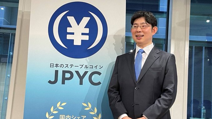 A man in a suit and glasses stands smiling beside a JPYC (Japan's Stablecoin) banner featuring the yen symbol, as Japan's Financial Services Agency moves to permit stablecoin transactions exceeding 1 million yen.