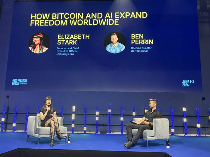 The image shows a stage with two people sitting down, possibly participating in an event or panel discussion about Bitcoin and AI. The backdrop features a blue screen displaying three different images of people's faces, likely representing the speakers at the event. The text "How Bitcoin and AI expand freedom worldwide" is displayed on this screen, indicating that the main topic of discussion might be related to the intersection between these two technologies and their impact on society.