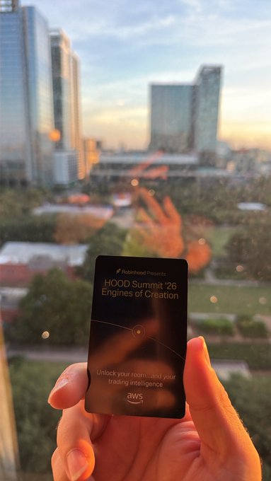 Hand holding an AWS-branded card for 'Hood Summit '26: Engines of Creation' with a city skyline blurred in the background.