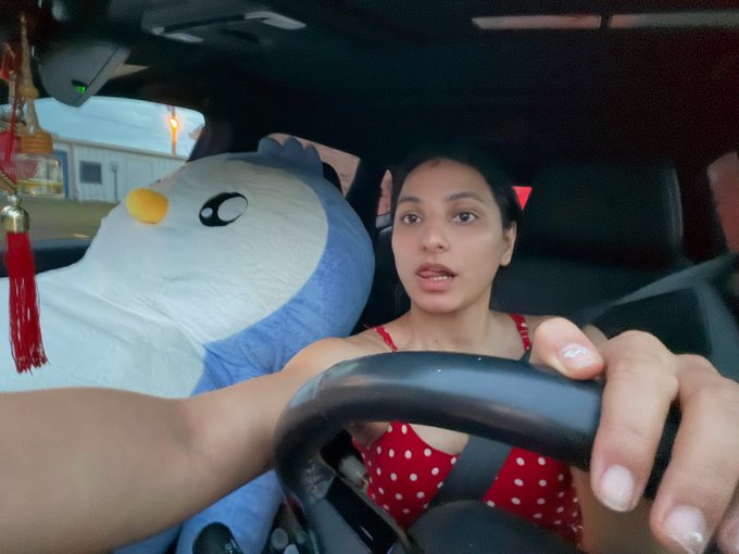 A young woman in a red polka-dot top drives a car with a giant plush penguin beside her, capturing the playful spirit of her "long-drive date with my giant pengu" for Pudgy Penguins' 5th birthday celebration.