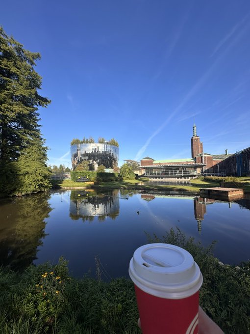 A hand holds a red coffee cup in the foreground, with a lake reflecting a modern circular building and a tall tower under a clear blue sky.