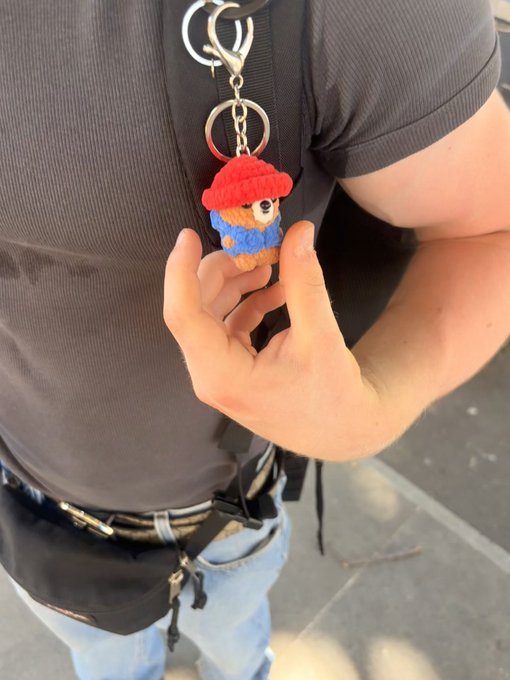 A person wearing a gray shirt and jeans holds up a small crocheted keychain charm depicting Paddington Bear, attached to a black bag strap.