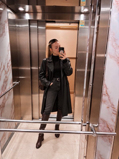 A mirror selfie of a woman in an elevator wearing a black leather trench coat, black top, gray jeans, and brown knee-high boots, with a small brown crossbody bag.