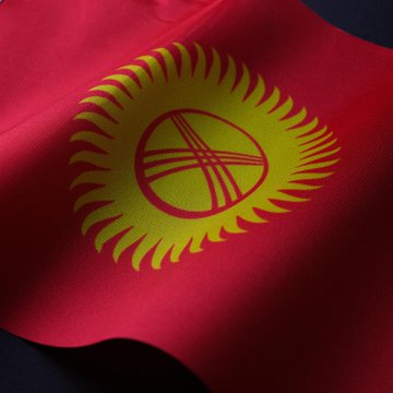 The flag of Kyrgyzstan waves against a dark background, its red field emblazoned with the iconic yellow sun and yurt-top symbol. The image accompanies news that the Kyrgyz government has ordered the wind-down of the gold-backed USDKG stablecoin, requiring all 50 million tokens to be redeemed or burned.
