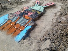 A colorful toy rabbit figure lying face-down on wet sand, possibly a craft project or kawaii art piece.