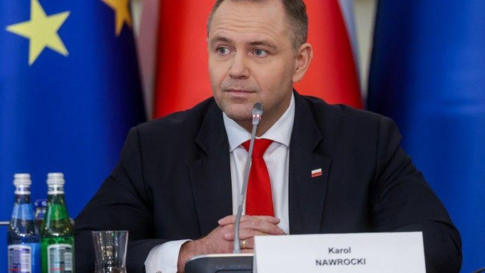 A photograph of Polish politician Karol Nawrocki seated at a table with a microphone, wearing a dark suit and red tie, with a nameplate reading 'Karol NAWROCKI' and flags visible in the background.