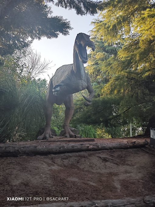 A large dinosaur statue, possibly a raptor, stands outdoors surrounded by trees, with a watermark 'XIAOMI 12T PRO | @SACRA97' visible at the bottom.