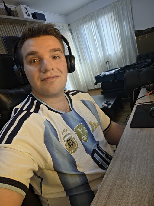 A man wearing an Argentina national football jersey and headphones takes a selfie while seated at a gaming/streaming desk setup.