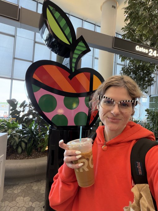 A smiling person wearing glasses and a red hoodie holds an iced coffee drink in an airport terminal, standing next to a large, colorful pop-art style apple sculpture near a 'Gates 24' sign.