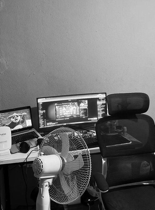 A black-and-white photo of a home office setup featuring a dual-monitor computer workstation, a standing fan, and an ergonomic office chair.