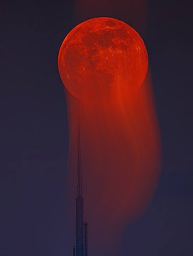 A striking nighttime photo of a large, blood-red full moon (likely a lunar eclipse) hanging low above a dark silhouette of a city skyline.