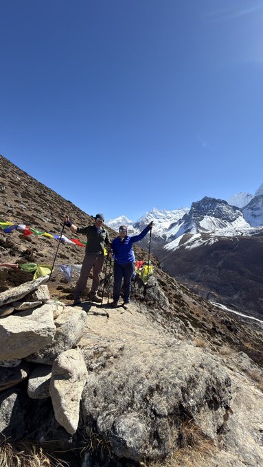 Two hikers pose with raised arms on a rocky mountain trail beneath a clear blue sky, with snow-covered peaks and prayer flags in the background.