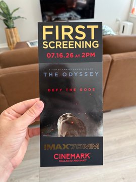 A hand holds a promotional ticket for the first screening of the film "The Odyssey," directed by Christopher Nolan, scheduled for 07.16.26 at 2 PM in IMAX 70mm. The ticket's date suggests the post "ONE YEAR LATER WE ARE SEEING THE ODYSSEY TODAY" is a humorous or speculative reference to the film's future release.