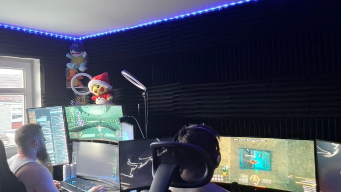 Two people wearing headsets sit at a gaming setup with multiple monitors displaying racing and simulation games, with plush toys stacked on a shelf in a soundproofed room.