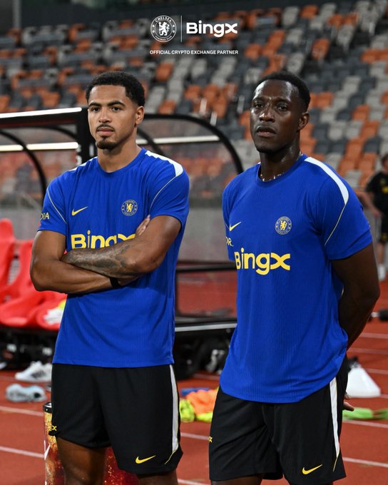 Two Chelsea football players stand on a track inside a stadium, wearing blue Chelsea training kits with the "BingX" sponsor logo across the chest and the club crest on the sleeve. The image features a "BingX" watermark at the top with "OFFICIAL CRYPTO EXCHANGE PARTNER" text, tying into the post's "#BingXCFC" branding and its theme of patiently waiting for the right moment to buy the dip, much like the focused players waiting on the pitch.