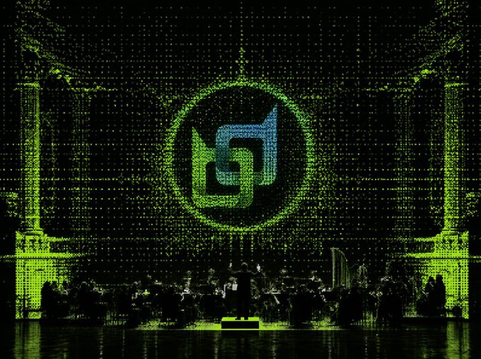 A stage performance with green lighting illuminating an orchestra, featuring a large projection of a stylized 'G' logo on the backdrop.
