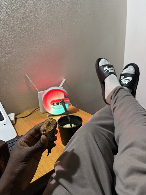 The image shows a person relaxing with their feet propped up on a desk, holding what appears to be a piece of food, with a colorful LED alarm clock displaying "10:32" in the background and Nike slides visible on their feet, illustrating a moment of affordable comfort and leisure within their last $5 budget.