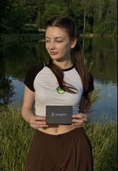 A young woman in a brown dress standing in a grassy field holding a small black 'Ledger' crypto hardware wallet device.