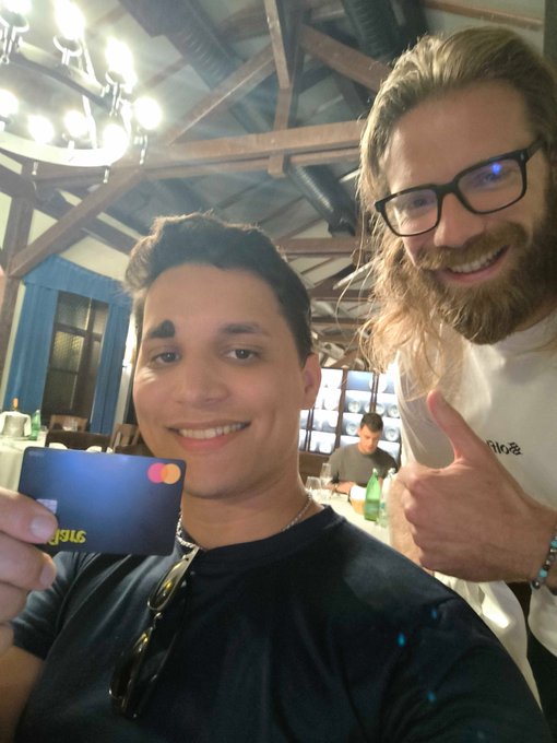 Two men smile for a selfie in a rustic restaurant with exposed wooden beams and a chandelier; one holds up a blue Mastercard with yellow text visible on the back, while the other gives a thumbs-up wearing glasses and a white shirt.